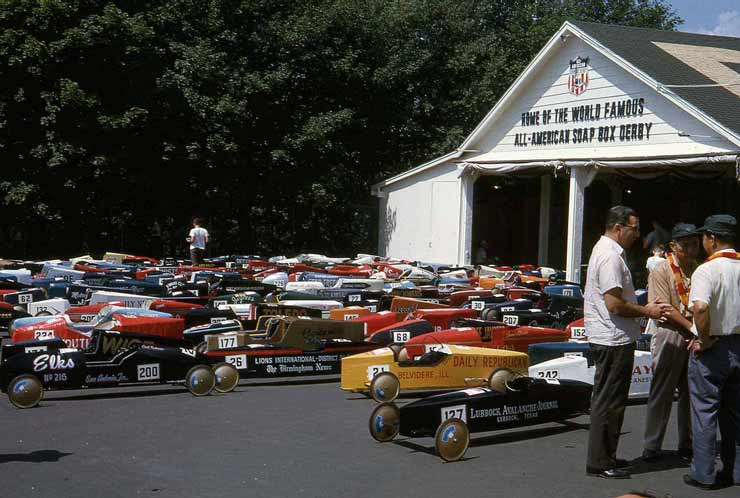 Over 50 multicolored soapbox derby cars from across the USA are parked in a sunny lot awaiting the race. Three men are chatting in the foreground and one man is walking through the cars in the background.